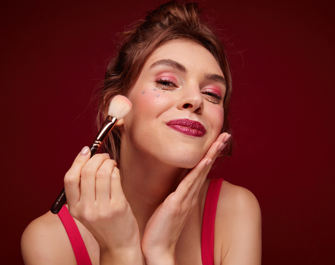 Mujer con labios rojos sonriendo con una brocha de maquillaje en su mejilla